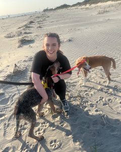 Corey Richardson on the beach with two dogs during a walk on the sand, smiling as one dog looks up at her.