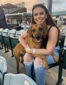 Corey Richardson smiling and holding a brown dog on her lap at a dog-friendly baseball game.