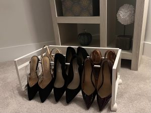 Compact white rolling shoe rack holding six pairs of women’s heels, neatly arranged by style and color, placed on a carpeted floor in front of a light wood shelving unit with decorative boxes and a faux flower display.