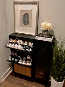 Modern black shoe cabinet with two pull-down shelves filled with sneakers, a decorative top surface featuring a framed abstract art piece, stacked books, and a vase with white flowers. A tall green plant in a white textured pot stands beside the cabinet, adding a natural touch to the corner of the room.