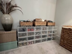 Stacked clear plastic shoe boxes organized in a corner of a carpeted room, each containing neatly stored shoes. On top of the boxes are woven baskets holding purses and accessories. The setup is flanked by a large ceramic vase with decorative grasses on the left and a woven laundry basket on the right, creating a warm and organized space